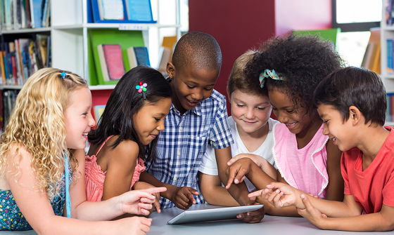 A group of children in a library gather around a tablet computer